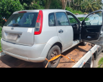 Foto de um carro da Ford cinza, com detalhes nas luminárias na cor vermelha, com a porta de passageiro aberta, colocado sobre um guincho. ao fundo uma área verde com várias árvores
