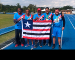 Foto de atletas do TRT do Maranhão com o treinador Emerson Viana. A foto foi feita numa pista de atletismo, localizada numa área verde. Os atletas e o técnico estão com camisas azuis e seguram a bandeira do Maranhão. Ao fundo, há um tenda branca, carros estacionados e uma casa.