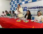 Imagem da juíza Liliana Bouéres proferindo discurso em pé com microfone ao centro da mesa do evento composta por autoridades que se encontram sentadas. A mesa está coberta por uma toalha vermelha e ao fundo tem um painel em tons branco e azul.