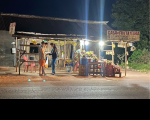 Imagem noturna de banca de frutas em rodovia onde se vê três pessoas.