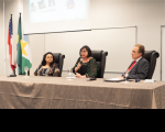 Foto da mesa de honra em primeiro plano, com toalha marrom claro. à mesa, duas mulheres e um homem, este no lado direito. A mulher ao centro, fala ao microfone. À esquerda, veem-se três bandeiras, dentre as quais a do Brasil no meio. No topo, uma tela de projeção mostra imagens não identificáveis.