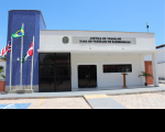 Foto da fachada principal da Vara do Trabalho de Barreirinhas, no Estado do Maranhão. No primeiro plano, entrada da VT com rampa e porta de vidro escuro. Paredes brancas e janelas de vidro escuro. À esquerda, coluna em lajotas azuis, à frente da qual veem-se as bandeiras do Brasil (centro), do Maranhão (esquerda) e do Município (direita). No topo, brasão do Poder Judiciário e, ao lado deste, as inscrições Justiça do Trabalho e Vara do Trabalho de Barreirinhas.