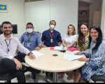 Foto de servidores reunidos no Setor de Ouvidoria do TRT do Maranhão, todos sentados ao redor de uma mesa redonda na cor bege, ao fundo se vê parede e armários em cor clara, e da esquerda para a direita os servidores Rafael (camisa branca e calça preta) André e Manoel (vestindo camisas em tons de azul e usando máscaras); e as servidoras Joelma (blusa branca), Vanessa (blusa amarela com detalhes pretos) e Edvânia (usando vestido em tons de azul)