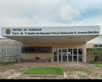 Foto da sede da Vara do Trabalho de Bacabal, antes da fachada principal há um canteiro com grama, na fachada também se vê portas de vidros de acesso à sede, além de outros canteiros na frente e plantas nas laterais, na parte superior da fachada se vê o brasão nacional e ao lado está escrito: Justiça do Trabalho - Vara do Trabalho de Bacabal - Fórum Raimundo B. Amaral Martins