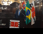 Foto vice-presidente do Coleouv e ouvidor do TRT-4, desembargador Clóvis Santos, que está discursando num púlpito com a logomarca do Tribunal de Justiça de São Paulo, atrás dele estão as bandeiras do Brasil (centro), São Paulo (à esquerda) e a do Tribunal de Justiça. No ambiente também há bancadas e armários em madeira escura, além de luminárias e detalhes da parede do Salão do Júri do TJ.