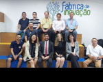 Onze pessoas sentadas em degraus e outra em cadeira, tendo ao fundo e no alto a representação gráfica de árvore compondo a marca do projeto Fábrica de Inovação.