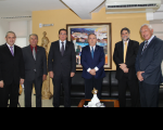 Cinco desembargadores do TRT e o ministro do TST em pé, lado a lado, posando para o registro fotográfico. Ao fundo, parede amarela, quadro com imagens de casas em espaço urbano.