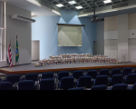 Foto com vista panorâmica do palco do Auditório Ari Rocha com os alimentos a serem doados dispostos ao centro. Bandeiras do Maranhão e do Brasil hasteadas à esquerda. Cadeiras azuis do Auditório na parte de baixo da foto. Telão de projeção de imagens no topo, sobre parede azul claro.