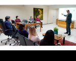 Foto no Gabinete do corregedor regional, desembargador Gerson de Oliveira, durante apresentação ao corregedor-geral da Justiça do Trabalho, ministro Caputo Bastos, equipe da Corregedoria. O desembargador Gerson está em pé, e o ministro e demais pessoas sentadas em cadeiras na cor cinza.  Sala na cor bege, onde se vê bancada e mesa de centro de madeira, mesa grande de madeira com tampo de vidro, um quadro pendurado na parede,
