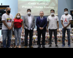 Seis pessoas em pé no palco de auditório tendo ao fundo telão da campanha Corregedoria Solidária e cestas de alimentos.