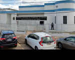 Foto da fachada principal da sede Vara do Trabalho de Timon. Sede pintada na cor branca, com detalhes na cor azul. O prédio é circundado por um gradil pintado na cor branca. Porta principal de vidro. Janelas também de vidro envoltas em gradil branco. Há um vigilante em pé na área externa da VT. Na fachada há o brasão do Poder Judiciário e ao lado está escrito: Tribunal Regional do Trabalho da 16ª Região - Vara do Trabalho de Timon. Há três carros estacionados na frente da vara trabalhista.