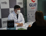 Foto de um homem e uma mulher: ele, médico oftalmologista, usando jaleco branco por cima de camisa branca com alguns detalhes e gravata preta; ela usando um blazer preto. Atrás, na parede, logomarcas do HRO e Geap.