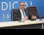 Foto do desembargador James Magno, usando óculos, terno escuro, camisa branca e gravata azul, gesticulando com ambas as mãos, sentado à mesa, falando ao microfone. Ao fundo, parede azul com parte de inscrição branca.