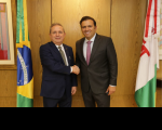 Foto de dois homens claros, vestindo ternos escuros e cumprimentando-se com aperto de mãos, ladeados por bandeiras e com parede amadeirada ao fundo.