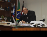 Foto com fundo branco e bandeiras do Maranhão, Brasil e da Justiça do Trabalho, tendo ao centro o presidente do TRT no Maranhão, desembargador Carvalho Neto, sentado em seu gabinete, vestindo terno escuro.