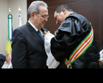 À esquerda, o presidente Carvalho Neto vestido de terno escuro recebe a medalha do desembargador Luis José de Jesus Ribeiro que usa toga e que se encontra à direita e de frente para o homenageado. Ao fundo duas bandeiras em mastros