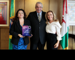 Foto de um homem grisalho, usando terno e gravata, entre duas mulheres, uma de cabelos escuros e outra de cabelos claros. Eles estão ladeados por bandeiras à frente de uma parede amadeirada.