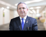 Foto do desembargador Carvalho Neto, usando terno azul escrito, camisa branca e gravata azul roial, com fundo desfocado claro, em tons de bege.