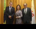 Foto de dois homens e uma mulher, em pé, à frente de parede amadeirada. Atrás estão duas bandeiras.