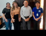 Foto de três mulheres e um homem que estão em pé, em frente a uma porta de madeira, numa sala onde se vê uma mesa, computadores e outros materiais.