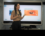 Foto de uma mulher jovem, de cabelos escuros e compridos, à frente de uma tela de apresentação de doenças oculares.