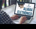 Foto de um notebook aberto sobre uma mesa, com uma pessoa digitando. Na tela, vê-se o texto ALVARÁ ELETRÔNICO em azul escuro e a logomarca do TRT-MA na versão colorida.
