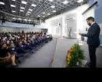 Foto de um homem em pé em um palco falando ao microfone e de uma plateia