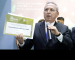 homem de terno, com microfone em um mão e na outra um certificado 