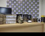 Coldres sobre mesa ao lado de pacotes e tela de computador. Ao fundo, painel com a logomarca da Polícia Rodoviária Federal.