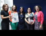 cinco mulheres posam para foto, uma das quais segura um cartaz do inovalab. Ao fundo, parede azul e janela com iluminação solar.