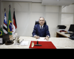 homem de terno sentado à mesa de gabinete. Ao fundo, bandeiras hasteadas. 