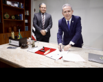 dois homens de terno posam para a foto, um dos quais assina um documento sobre a mesa