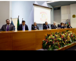 mesa de honra com oito homens sentados. Arranjo de flores na frente da mesa. Ao fundo, imagem de  data show e bandeiras hasteadas.