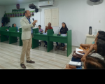 Foto de um homem branco em pé numa sala de paredes brancas, falando ao microfone, rodeado de pessoas sentadas em carteiras escolares verdes.
