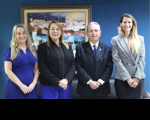 Foto de três mulheres e um homem com as mãos cruzadas à frente de seus corpos. Ao fundo, parede azul marinho com um quadro de arte colorida.