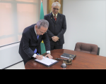 Foto de dois homens usando ternos escuros. Um deles está reclinado sobre uma mesa, assinando um documento. O outro está de pé logo atrás, observando a assinatura.