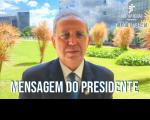 Ao fundo, gramado e plantas que decoram um jardim. À frente, homem vestido com terno e gravata em tons escuros, usando óculos de lentes arredondadas. Texto escrito de cor branca. 