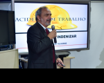 Foto de homem branco com cabelos e barba grisalhos, de perfil, usando terno escuro e falando ao microfone, na frente de uma tela de datashow mostrando um capacete amarelo e o texto ACIDENTE DE TRABALHO em letras azuis.