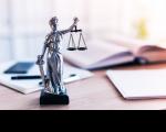 Estatua da Justiça sobre a mesa. Ao fundo, estão desfocados um livro aberto, papeis de anotação e uma caneta