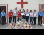 Parede de fundo na cor branca e uma cruz centralizada na cor vermelha, porta de madeira com grades ao lado esquerdo. Nove pessoas, homens e mulheres, posando para a foto com o cartaz da campanha em mãos. À frente, alimentos arrecadados.