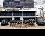 Policiais judiciais fardados, posam para foto em frente a fachada do prédio-sede do TRT-16, ladeados por duas viaturas pretas. O grupo esta perfilado em "V", tendo à frente uma mulher e apenas homens nas laterais.
