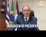 Homem sentado em uma cadeira preta, usa óculos de grau com lentes arredondadas, um terno azul escuro, uma camisa branca e uma gravata azul. Ao fundo, uma parede na cor branca, três bandeiras hasteadas e uma estante com livros à esquerda.