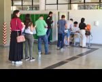 Fila com candidatas e e candidatos para entrar na sala de aplicação de prova. Homem sentado em carteira coletando as informações.