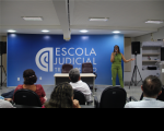 Ao fundo, uma mulher vestida com um macacão na cor verde e falando ao microfone. Ao fundo, uma caixa de som preta e uma parede azul com letras brancas, com texto escrito ESCOLA JUDICIAL. À frente, pessoas sentadas em cadeiras pretas. 