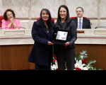 Duas mulheres posando para foto, com vestimentas formais, segurando uma placa de premiação. Atrás delas um arranjo central de flores vermelhas e brancas. Ao fundo,  uma mulher e um homem estão sentados em poltronas vermelhas, apoiados em uma mesa.