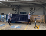 A imagem mostra dois homens usando equipamento de proteção individual, carregando painel solar em formato retangular, de cor azul escuro. À direita, um equipamento de transporte das placas solares. Ao fundo, placas empilhadas sobre pallet de madeira. 