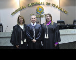 Foto de um homem claro, ladeado por duas mulheres de cabelos claros. Todos usando togas de juiz e posando para a câmera fotográfica. Atrás deles, parede com a inscrição TRIBUNAL REGIONAL DO TRABALHO em letras douradas, acima de um brasão colorido da República do Brasil.