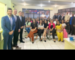 Grupo de pessoas posando para foto, alguns na parte central, sentados em cadeiras e outros, dos extremos, em pé. A foto acontece dentro de uma sala com paredes na cor branca.