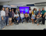 Grupo de pessoas, posando para foto, alguns na parte central sentados em cadeiras e outros em pé. Ao fundo, vê-se tela de projeção em cor predominantemente roxa, com texto na cor branca escrito "PROGRAMA TRT NA ESCOLA 2023". A foto acontece dentro de uma sala com paredes na cor branca.