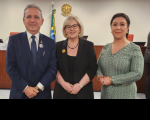Três pessoas posando para foto, sendo um homem e duas mulheres. Ambos estão com as mão cruzadas à frente de seus corpos. O homem, ao lado esquerdo, vestido com terno azul escuro, a mulher que está no meio está vestida com roupas de cor preta, e a da esquerda está com um vestido verde estampado.